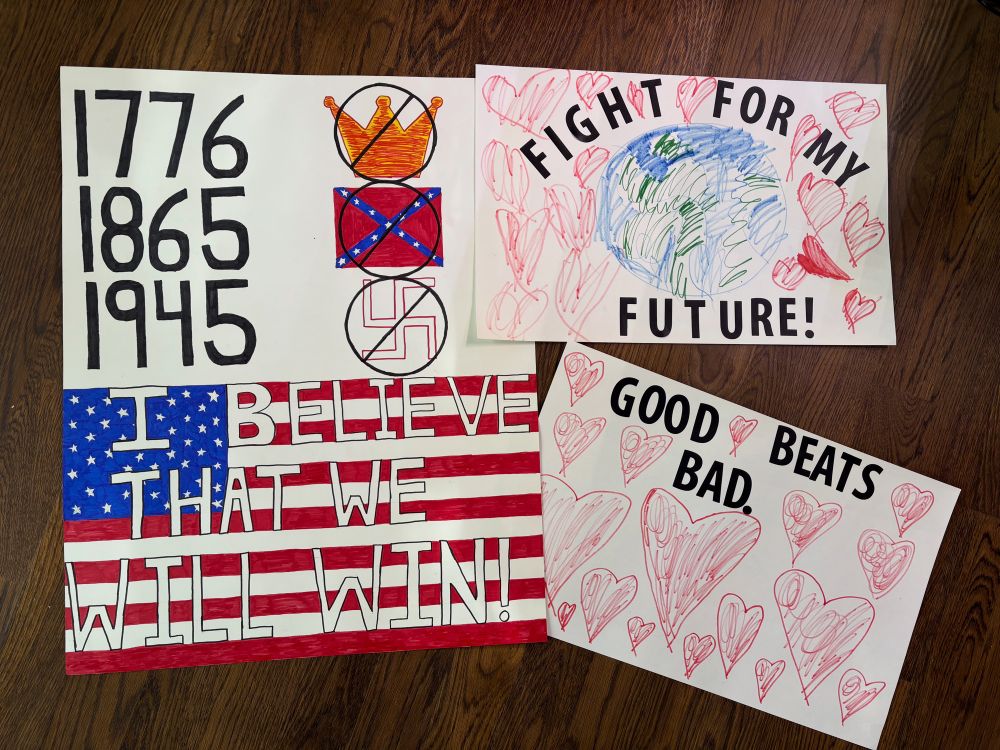 Three protest signs. One says “I Believe That We Will Win” over the American flag with important dates marking America’s Declaration of Independence in 1776, the fall of the confederacy in 1865, and the defeat of the Nazis in 1945. Another says “Fight for my Future” around a child’s hand drawn picture of planet Earth. The third sign says “Good beats Bad.” And is surrounded by a child’s drawing of many hearts.