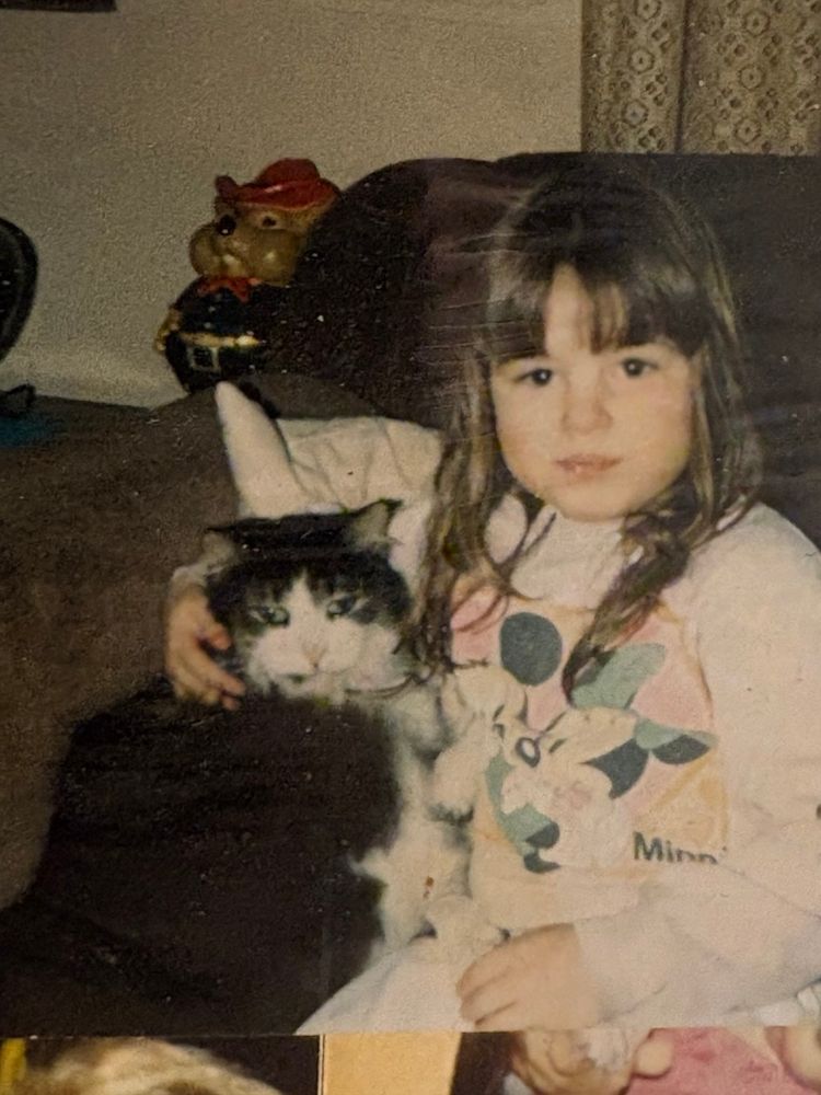 A young brunette girl with bangs and long hair. Shes wearing a long sleeved micky and Minnie Mouse shirt with her arm around a scraggly old girl cat whose eyes are looking in different directions. Everything is cigarette yellow lol. Probably 1990? 