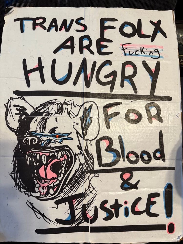 A white and black sign. The background is white and the drawing is in black. There’s a drawing of a hyena with its fangs bared and a snarl. It has pink and blue highlights. There is also words that say Trans folx are fucking hungry for blood & justice! 