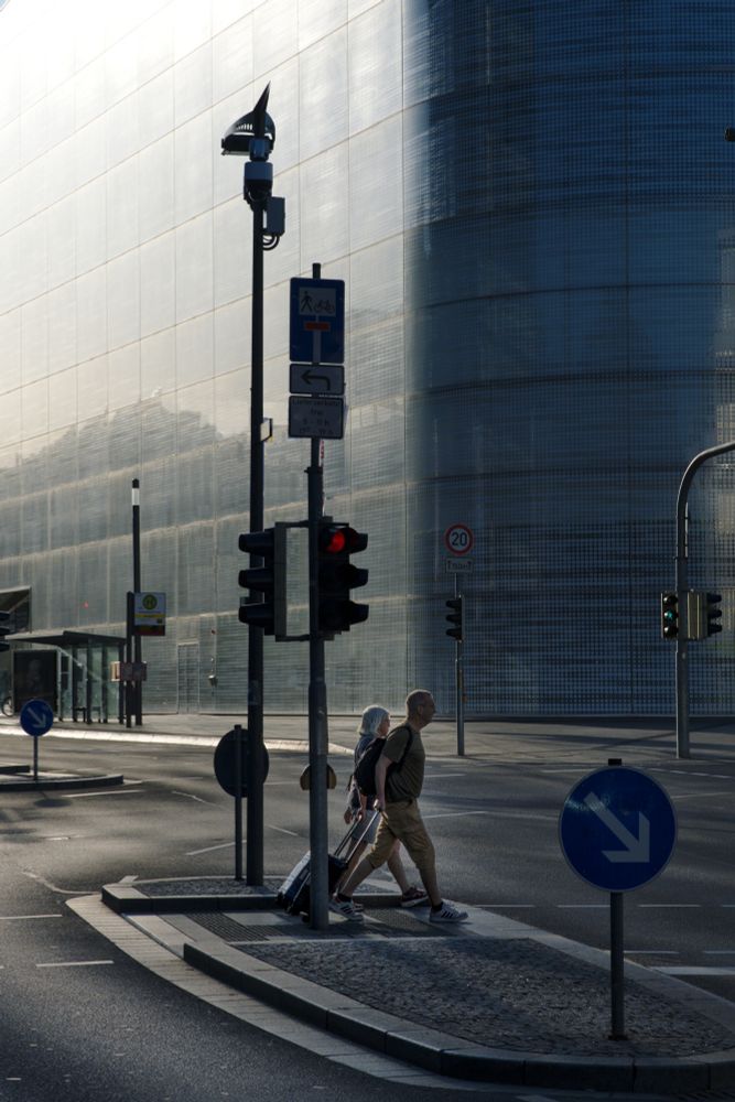 Two persons crossing a street in front of a futuristic building reflecting the autumn sunlight. Street lights and a CCTV tower structure the scene. 