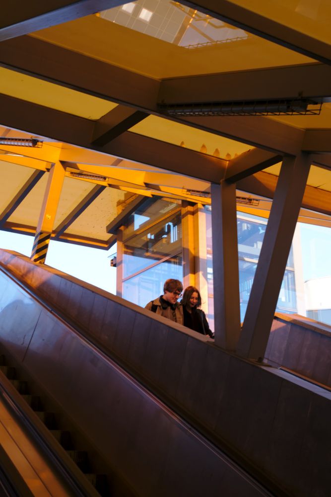 Two persons on an escalator down, in the orange light. 