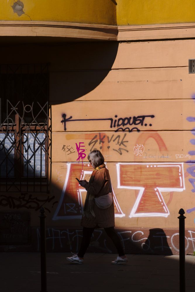 Woman walking along a graffitied orange housefront on which the sun casts sharp shadows; she is walking into a diagonal shadow, only her upper body is still in the light. 