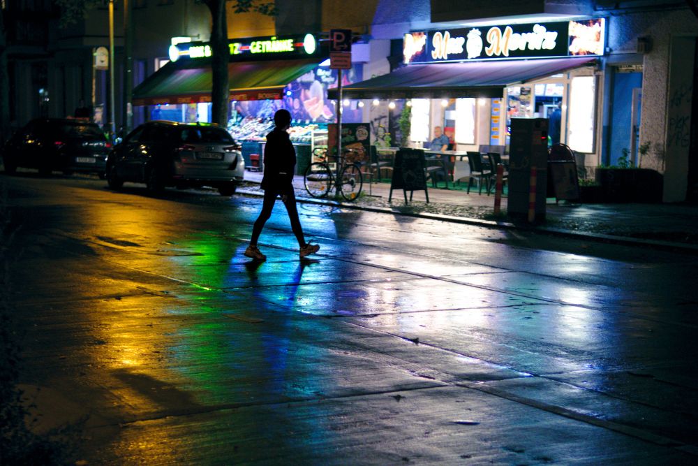 A dark figure crossing the street at night, some colorful reflections are visible on the wet surface. 
