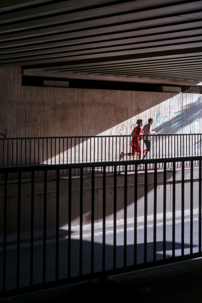 Two runners in an underpass, the sun casting diagonal shadows, railings with vertical bars structure the frame. 