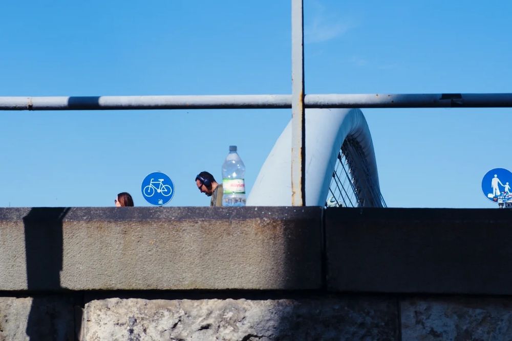 Two walkers, a bottle, two signs and a bridge behind a wall. The blue sky above. 