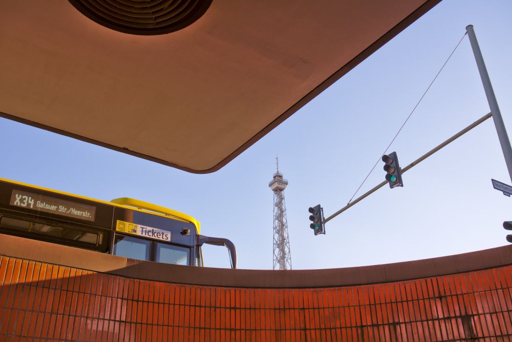 Photo of Berlin's old TV tower ("Funkturm"), taken from the exit of a nearby subway, so the tower is only half visible. The orange tiles and the modernist roof of the subway exit as well as a yellow budls and a traffic light frame the scene. 
