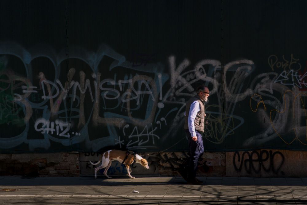 The sun illuminates a man walking his dog in front of a graffitied wall in the shade. 