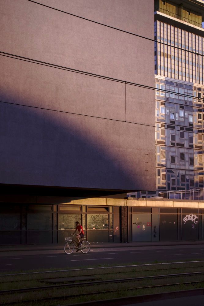 Bicyclist in front of large building in the evening sun. Shadow and light play on an overhanging simple facade. In the background, windows of an office tower.