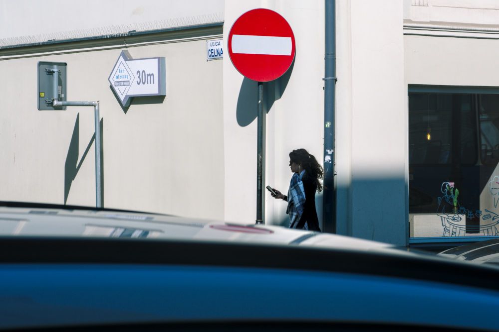 A street corner with white walls in the sun, a woman, a stop sign, all behind a shiny black car. 