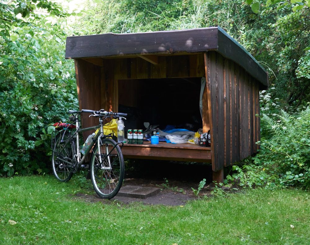 A camping box amidst verdant foliage, touring bike, camp gear and three cans of Pilsner Urquell 
