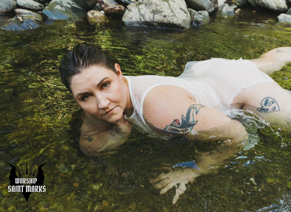Saint Marks lays in a tranquil river, her hair slicked back and translucent white dress draped across her skin. She gazes at the viewer hungrily.