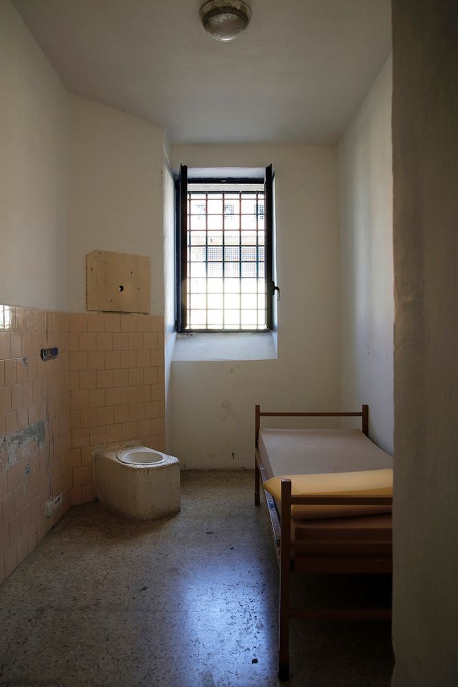 Imagem de um quarto estreito, com uma cama de madeira e um colchão tão fino quanto o travesseiro. Não há roupa de cama. Ao lado da cama uma privada sem tampa colocada num quadrado de concreto. Uma janela alta com grades está ao fundo. A cela tem azulejos velhos e alguns quebrados, do lado esquerdo da parede. Deve ser pior que o airbnb que ela alugou com dinheiro de minion trouxa.