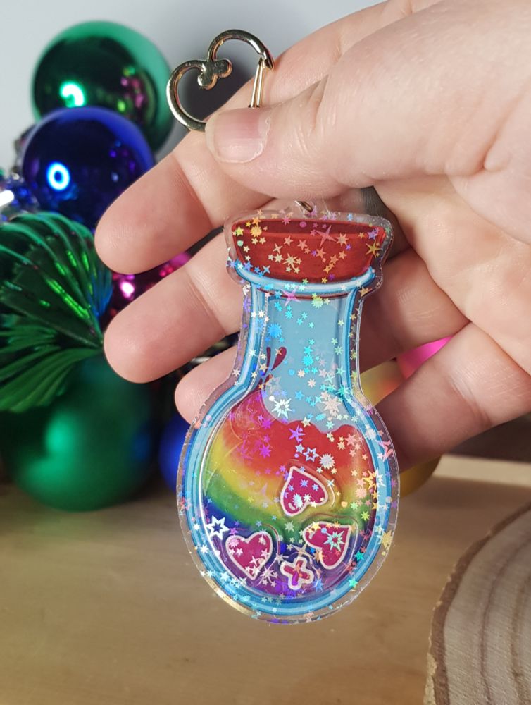 An acrylic shaker keychain of a rainbow coloured potion with shakeable red hearts in it.