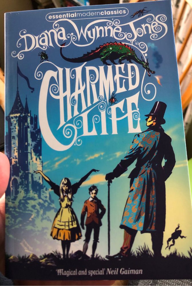Front cover of a paperback of Diana Wynne Jones’s ‘Charmed Life’. 
The picture is of a man, his back to us, facing a boy and girl. All in possibly Edwardian style dress. The girl’s arms are raised as if casting a spell. There’s a fantasy-style castle in the background and a dragon perched on the book’s title.