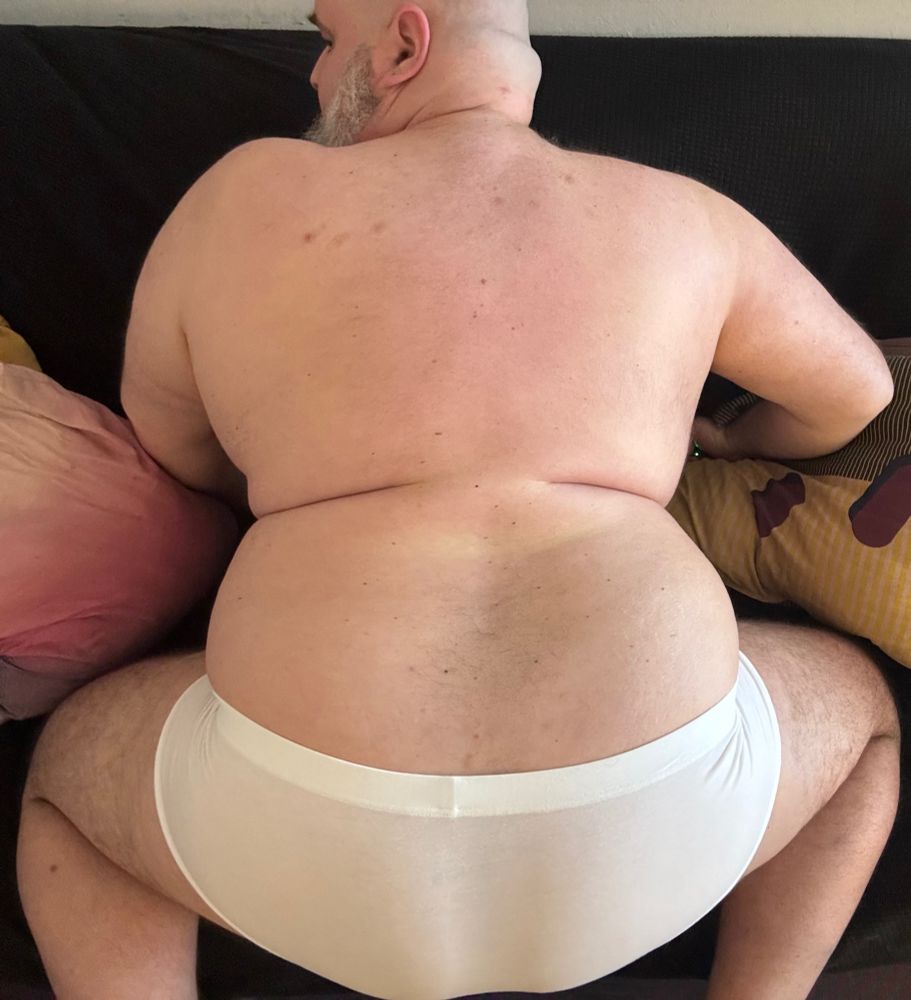 Top down shot of a fat guy in white briefs kneeing on a sofa. His head is turned to the left so you can see his salt and pepper beard. The photo is framed in a way that centers his broad back but cuts of the top of his head and everything below the ankles. No free feet pics.