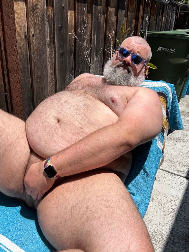 A fat man with a grey beard laying out in the sun wearing purple sunglasses.
He is shot from below while lying back on a chaise lounge and tastefully covering the goods with one hand