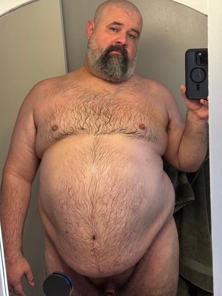 A picture of naked fat man with a shaved head and a salt and pepper beard taking a mirror selfie in his bathroom. He is wet having just gotten out of the shower. His large body fills most of the mirror centering on his big belly in keeping with the customs and traditions of tummy Tuesday