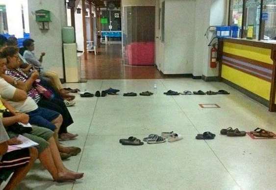 Foto que mostra diversas pessoas sentadas aguardando atendimento enquanto seus próprios chinelos guardam seus lugares na fila.