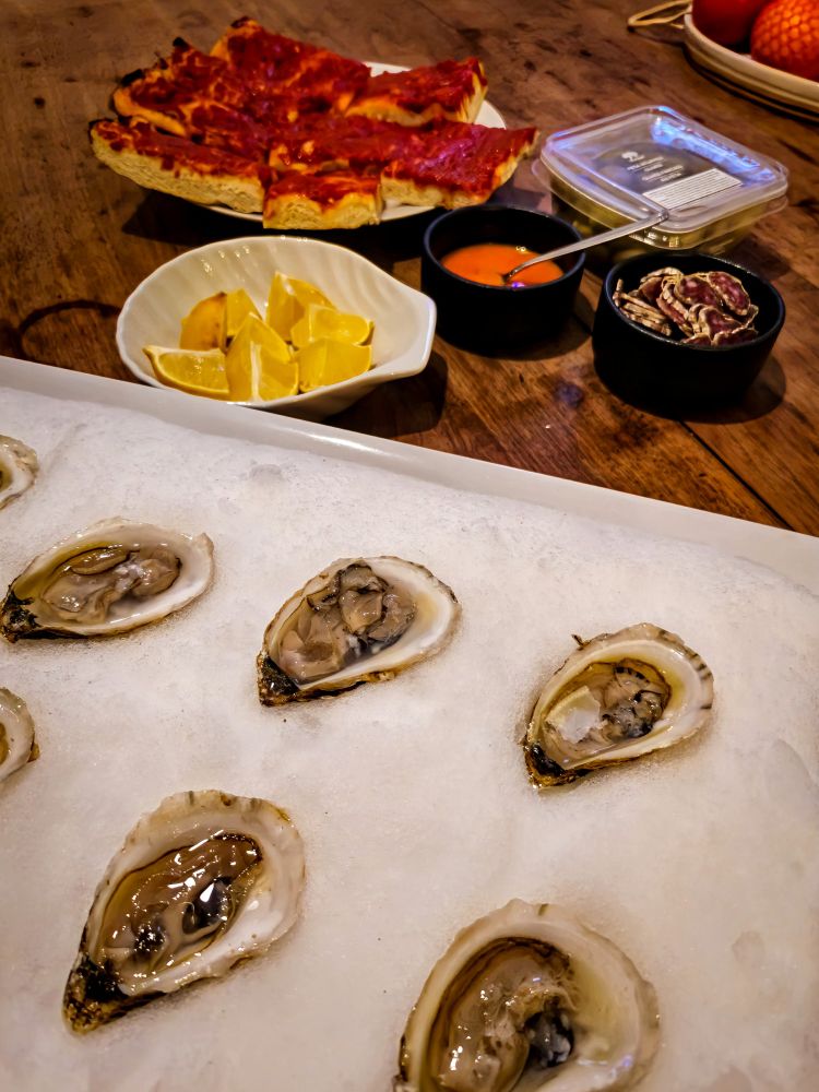 Table with oysters on snow, some lemons, tomato pizza and some sliced saucisson. 