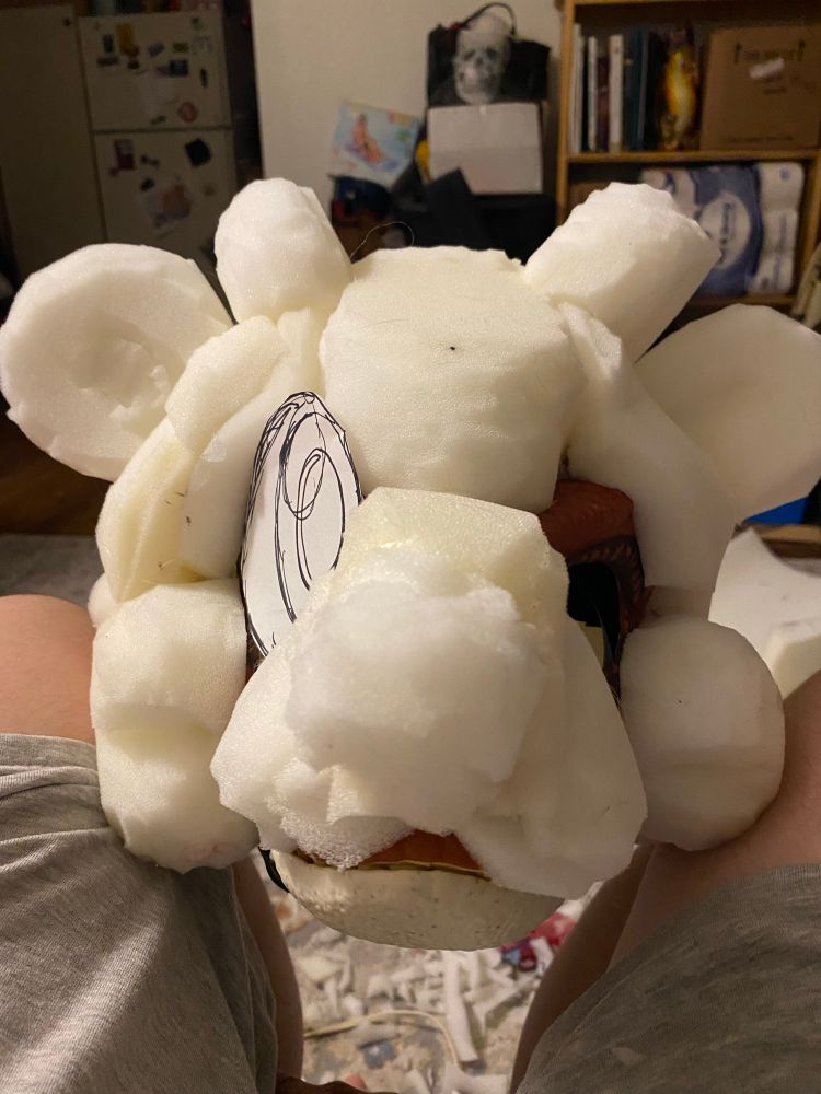 Front view of an in progress fursuit on a dino mask headbase. A paper eye template rests in the left eye hole
