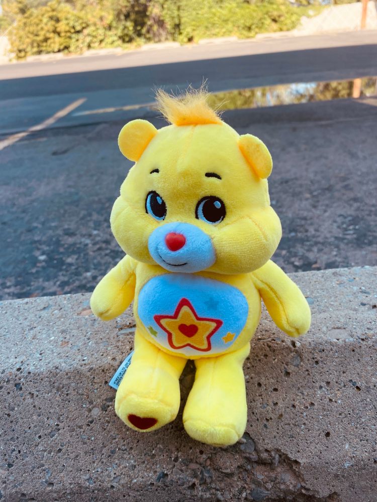 A picture of a bright yellow care bear (superstar bear) plush sitting on a cement parking brick