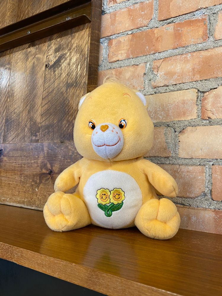 A Friend Bear Care Bear plush sitting on a wooden wall runner with a brick wall behind her