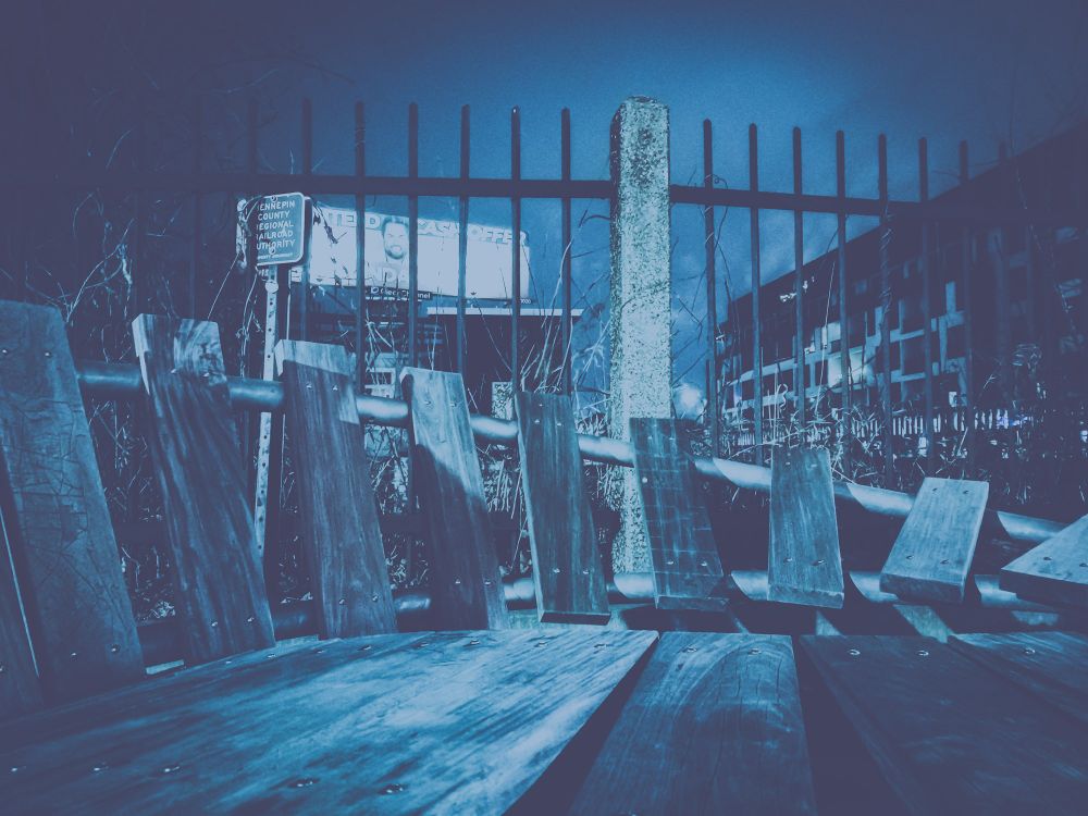 A wooden brutalist bench, in front of a real estate billboard, iron fence, and a luxury apartments at night, with an overwhelming blue 