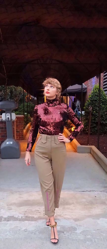 Taylor in a sparkly maroon top and high waisted brown pants, looking up at the sky with a curious expression, her wavy hair is pulled up almost in a bun and her left hand is on her hip