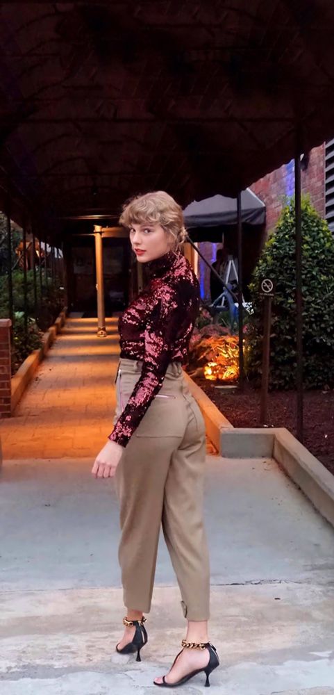 Taylor in a sparkly maroon top and high waisted brown pants, turning to face the camera with a curious expression, her wavy hair is pulled up almost in a bun