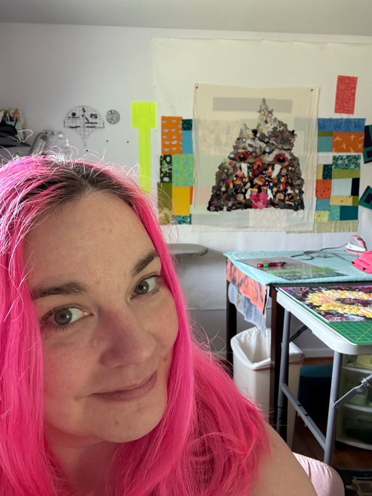 phone selfie of me, a white woman with pink hair, in a sewing room. behind me is a collage quilt of a mountain shaped hell mouth with red eyes and pointy teeth.