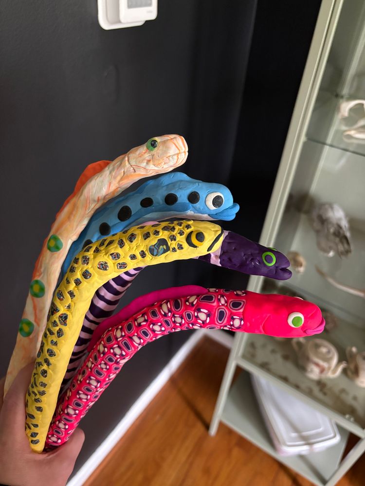 five doofy bright-colored polymer clay eels. inspired by garden eels. they are orange and white, blue with black spots, yellow with blue spots, purple/light purple striped, and hot pink with purple and white spots.