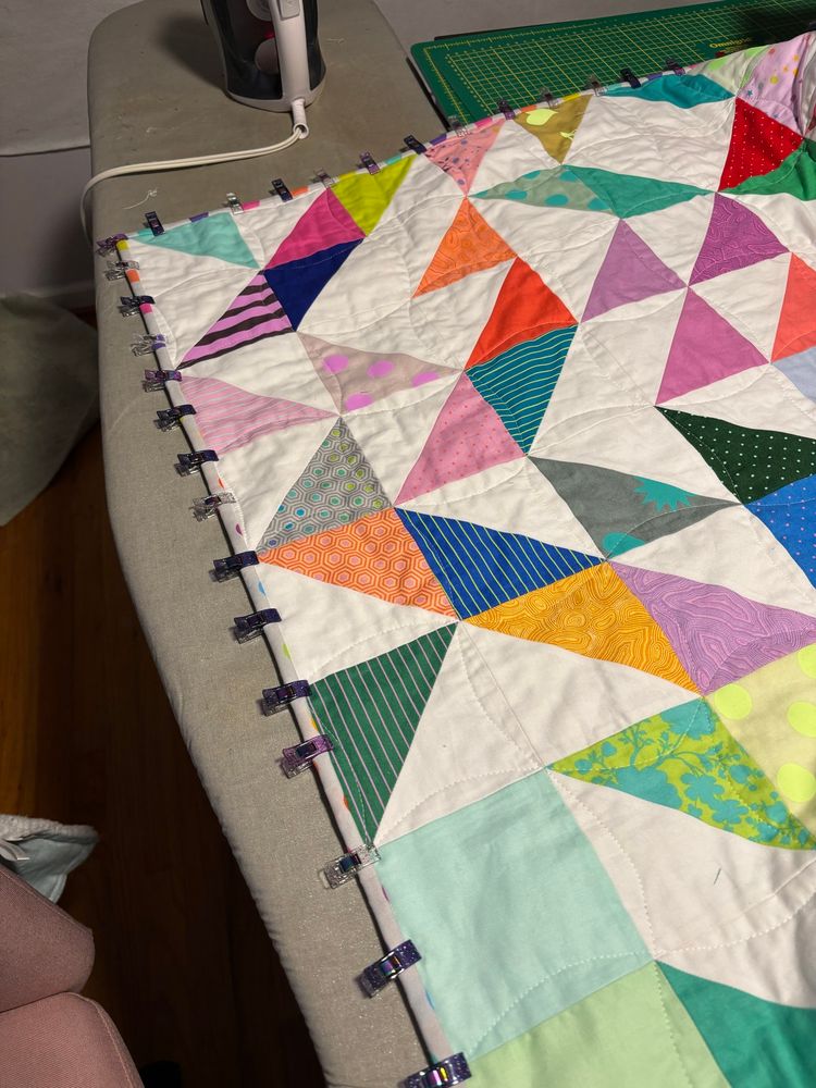 the corner of a quilt made of colorful triangles and squares with the binding held in place by little plastic clips