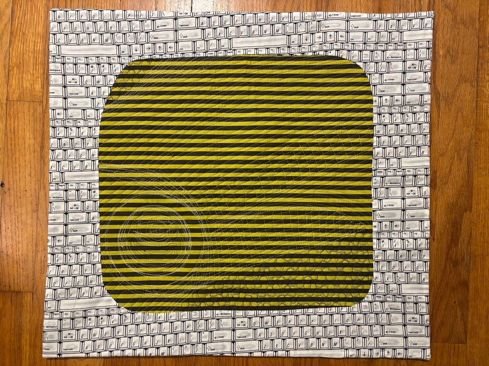 the back of the quilt. the quilting shows through on the green and grey horizontal striped backing. it’s framed by a white print of a computer keyboard.