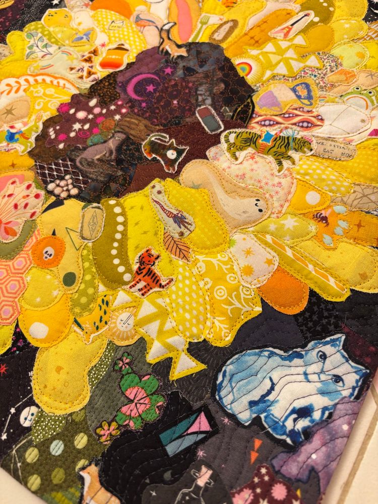 a collage quilt of a sunflower made up of hundreds of tiny pieces of fabric. the background is mostly grey, black, and purple prints. the petals are in the yellow family. the center is brown and purple. there’s a little bit of green stem showing. prints include foxes, otters, cats, an octopus, stars, skulls, leaves, and fruit.