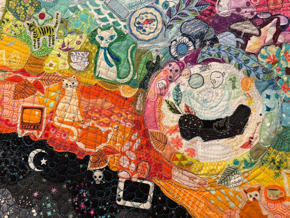 closer in on the eye of the squid. the eye is quilted with iridescent thread. the circle outside the eye is unquilted and puffy. some fabric motifs here are cats, flowers, a tv, ghosts, mugs, plates, gems, and mushrooms.