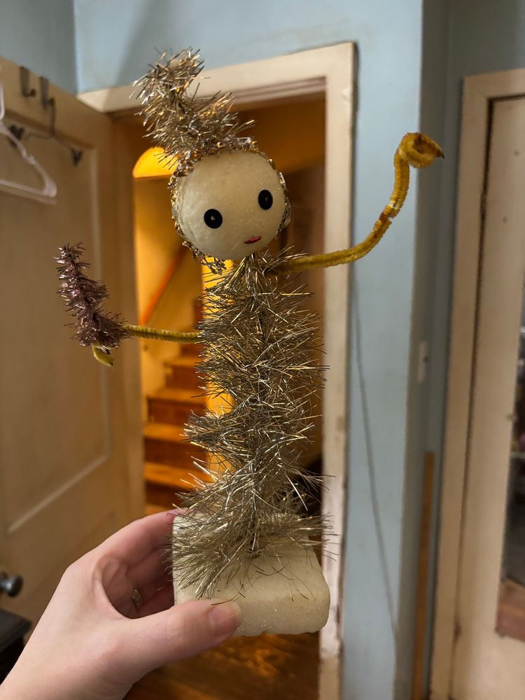 a very homemade christmas decoration of a styrofoam ball on top of some tinsel garland with pipe cleanser arms. the ball has a face of sequins and a little tinsel hat. it is holding a tiny tinsel tree