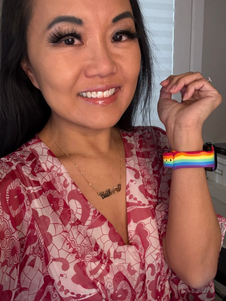 Selfie by Catherine Lewis. She is a Chinese Canadian woman with long black hair, wearing a flowered pink dress and a rainbow Pride Apple Watch band.