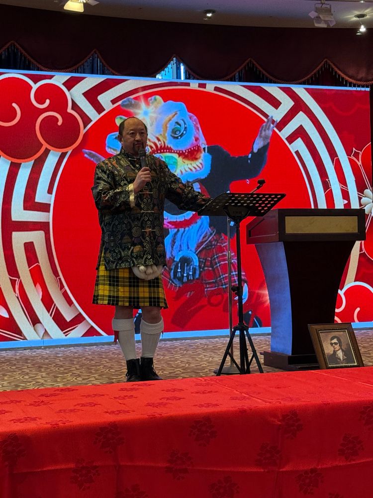 Event organizer Todd Wong on stage in a traditional Chinese jacket and a kilt
