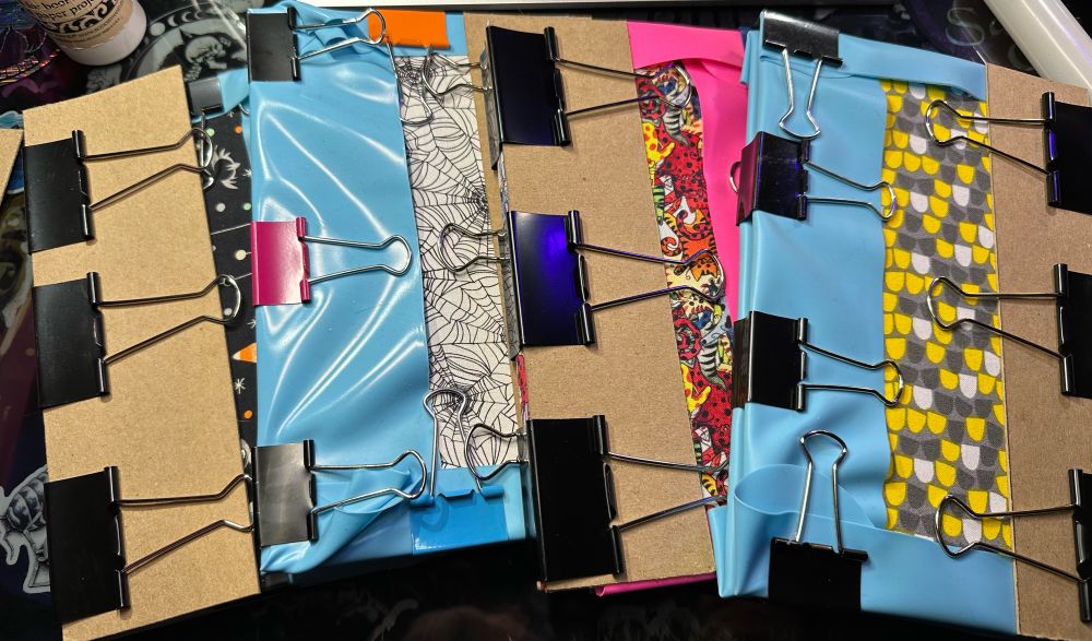 Four books wrapped up in binder clips and silicone shields to protect the inside from glue.
One is halloween treats on a black background, black spider webs on a white background, many many colorful cats on the next, and the last… well, I think it looks like decorative corn.  But that’s me.