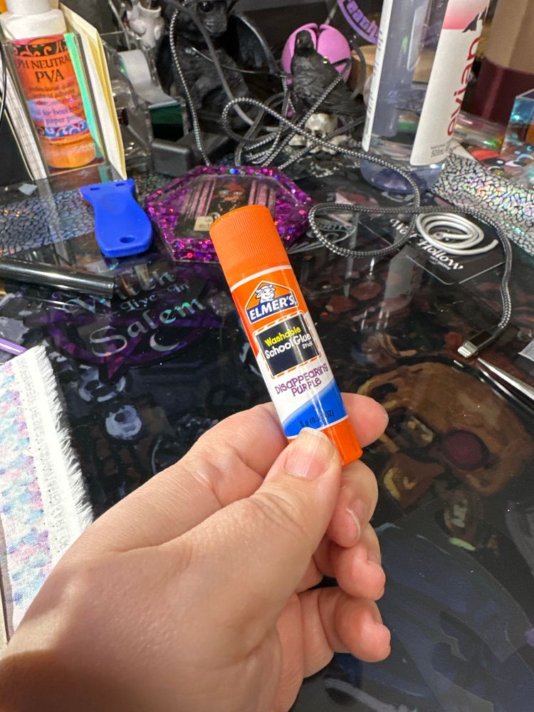 A stick of Elmer’s school glue being held in my hand with my work desk in the background. 