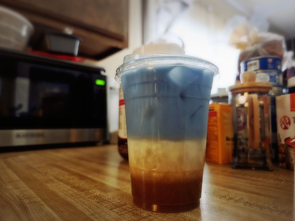 A layered coffee drink on a kitchen counter. The drink has blue liquid, oatmilk, and 3 espresso shots 