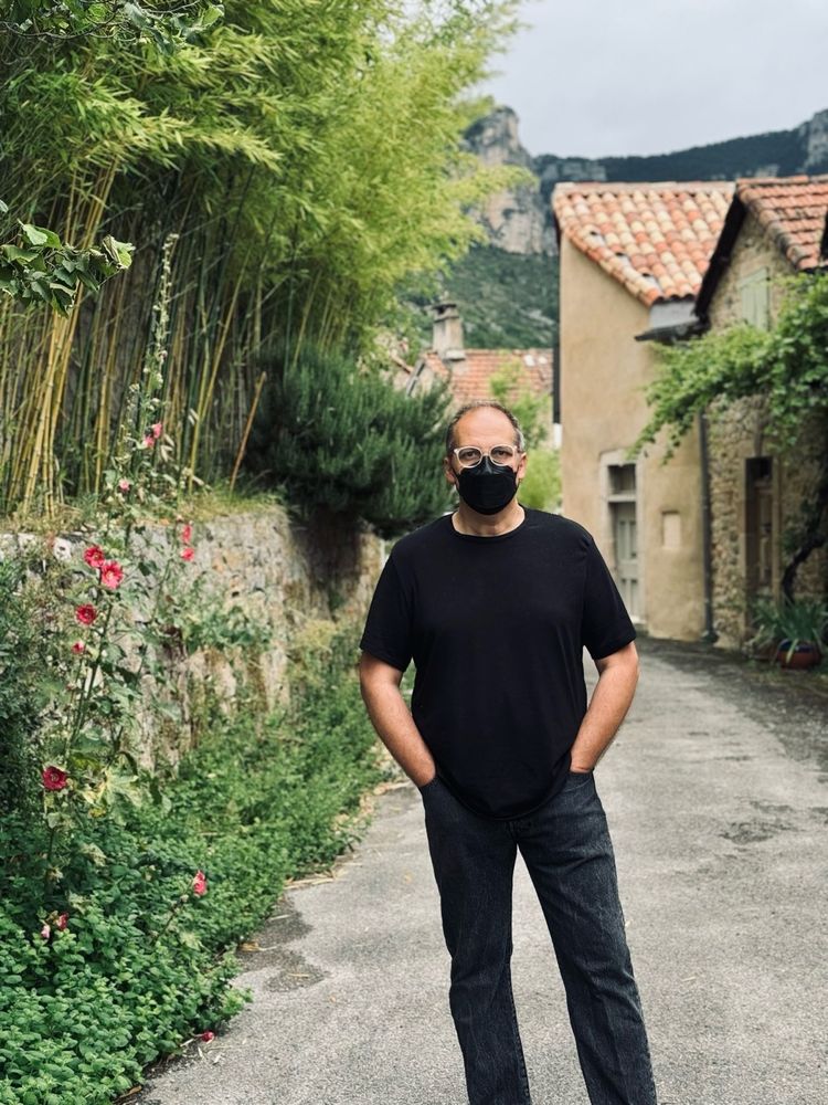 Selfie dans ma rue, vieux village, des maisons en pierre à l’arrière plan, des bambous sur le côté. Je porte un jean anthracite, un T-shirt noir, un FFP2 noir et des lunettes blanches.