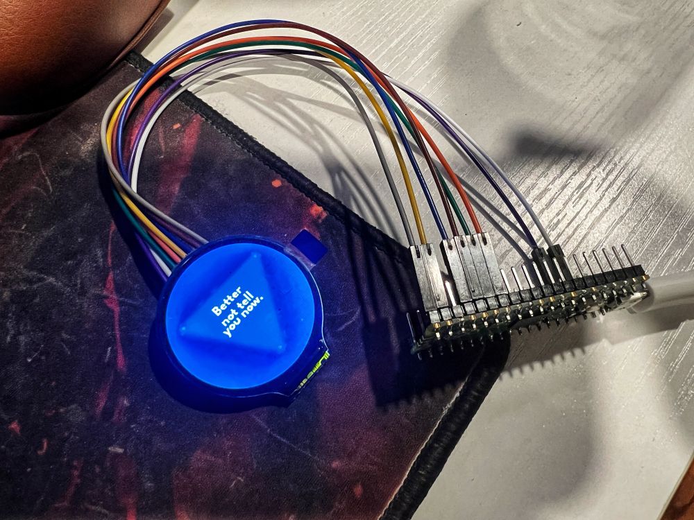 A circular display connected to a raspberry pi pico showing a blue triangle and the words “better not tell you now”.
