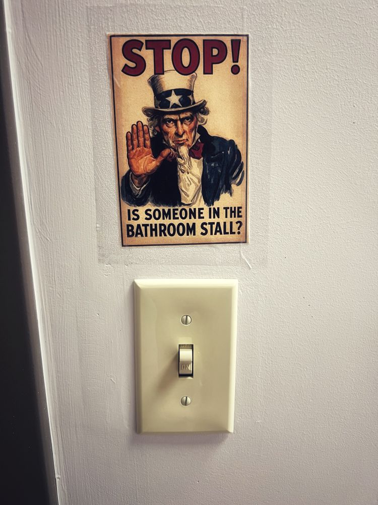 A world war 2 propaganda style picture taped above a bathroom light switch. It reads: STOP! Is someone in the bathroom stall?
