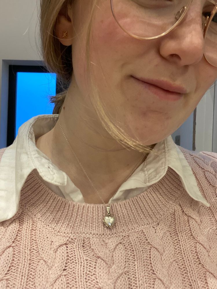 River is showing off her earrings which are tiny four pointed stars, wearing a pink knit jumper and a white shirt. You can also see her locket around her neck and her hair is pulled back with a strand of hair dangling down at the front.