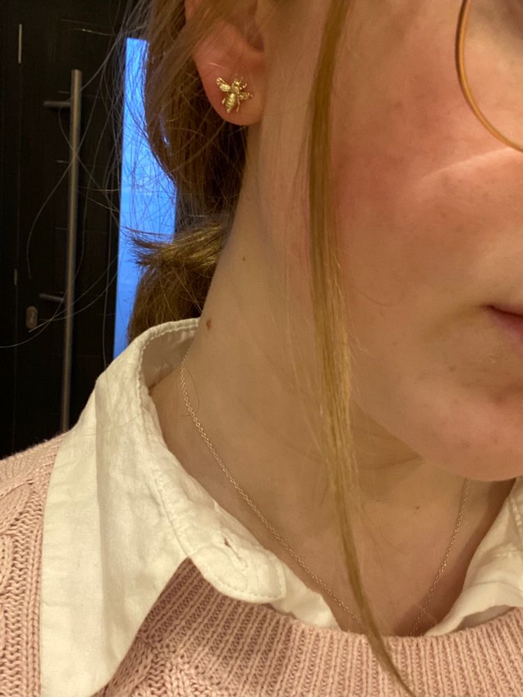 River is showing off her earrings which are tiny gold bees, wearing a pink knit jumper and a white shirt. Her hair is pulled back with a strand of hair dangling down at the front.