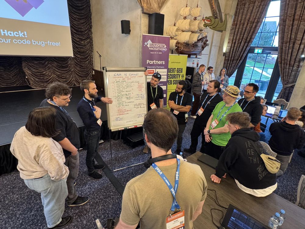 A group of people gathers around a presenter who is explaining ideas on a whiteboard at a hackathon event. The presenter, a bearded man in a dark shirt, gestures towards the board, which contains handwritten notes under the title “CVE IS COMING! Securing the Supply Chain.” Participants wear conference badges and lanyards, with some taking notes and others listening attentively. The venue has a high ceiling, decorative elements including a wooden ship mounted on the wall, and large windows letting in natural light. Banners in the background display event branding, with text such as “CloudFest Hackathon” and “Next-Gen Security for Open Source.” Tables with laptops and event materials are also visible in the room.