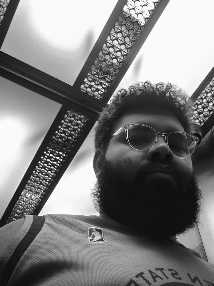 A imagem em preto e branco mostra um homem (eu) de barba e óculos, fotografado de baixo para cima. Ele está em um elevador e sua expressão é séria