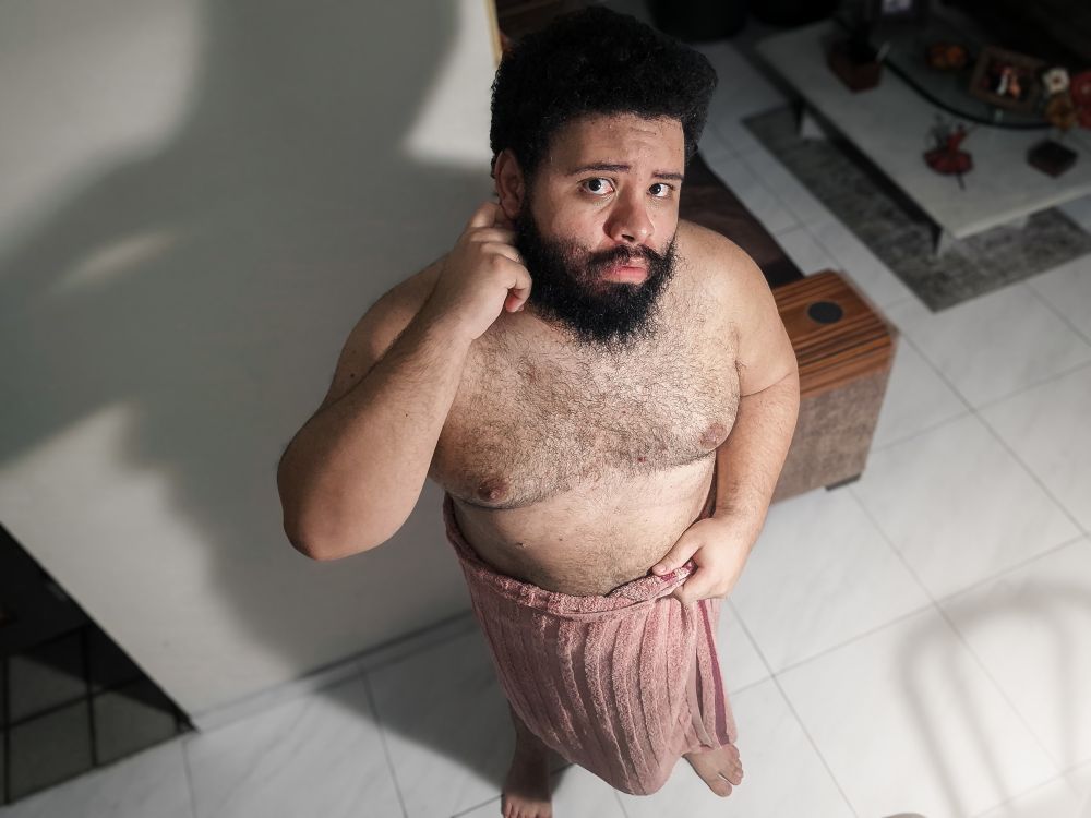 A imagem mostra um homem (eu) de barba e cabelo cacheado olhando para cima, com expressão pensativa. Ele está enrolado em uma toalha da cor salmão e posa em um ambiente interno com piso de cerâmica branca e uma sala ao fundo. A foto é tirada de cima, com iluminação que projeta sua sombra na parede