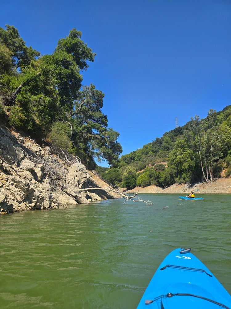 The blue nose of a kayak in aqua water. Another blue kayak in the distance. Green trees cover two hills on either side of the water. 